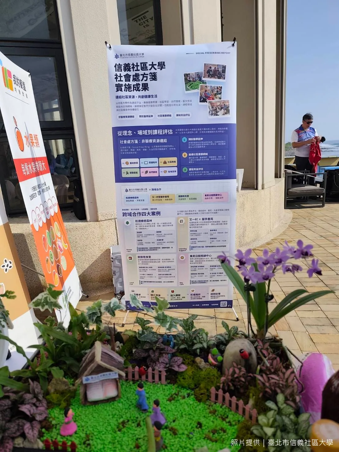 「信義社區大學社會處方箋實施成果」展板立在海邊活動現場，前景是課程的微景觀模型。